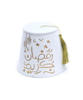 A white porcelain jar with a fez (Tarboush) shape, featuring hand-painted gold text 'Ramadan Kareem' and decorative elements, accompanied by a gold tassel.