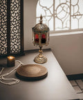 A metal lantern with multicolored glass windows, displaying a candle inside, placed on a table with a decorative tassel in the foreground.